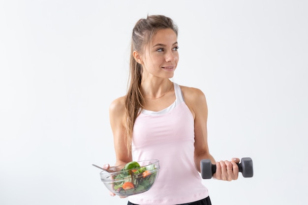 Estilo de vida saludable, concepto de personas: un plato de ensalada en una mano y una pesa en la otra. Sonrisa y traje de fitness.