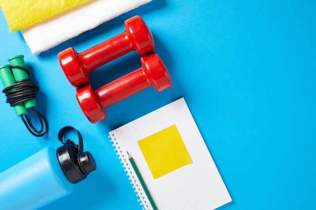 Foto estilo de vida saludable, perder peso y concepto deportivo. equipo del atleta: mancuerna roja, botellas de agua deportiva, cuaderno en blanco, bolígrafo, comba sobre fondo azul. espacio para texto. vista plana, vista superior