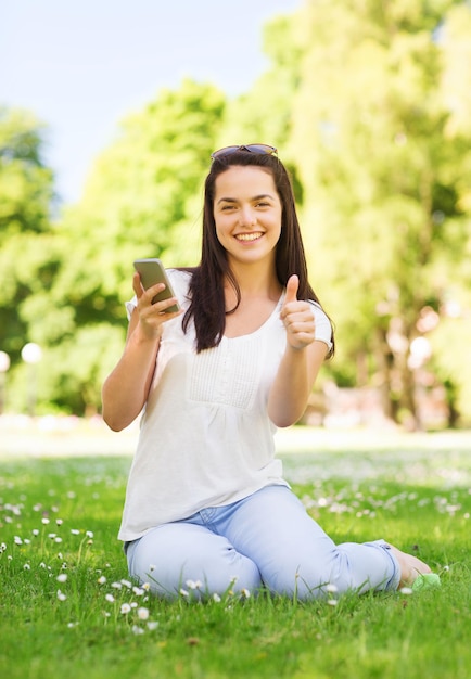 estilo de vida, vacaciones de verano, gesto, tecnología y concepto de personas - joven sonriente con teléfono inteligente mostrando los pulgares hacia arriba en el parque