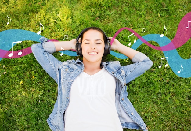 Foto estilo de vida, vacaciones de verano, tecnología, música y concepto de personas - jovencita sonriente con auriculares tendida con los ojos cerrados sobre el césped