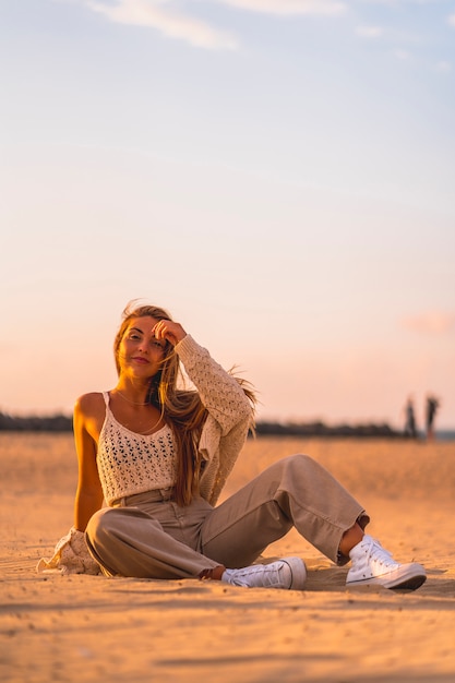 Estilo de vida de verano, una joven rubia con el pelo liso, con un pequeño suéter de lana y pantalones de pana sentada en la playa. Disfrutando de una tarde de verano al atardecer sentado en la arena