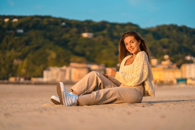Estilo de vida de verano, una joven rubia con el pelo liso, con un pequeño suéter de lana y pantalones de pana sentada en la playa. Sonriendo mucho al atardecer