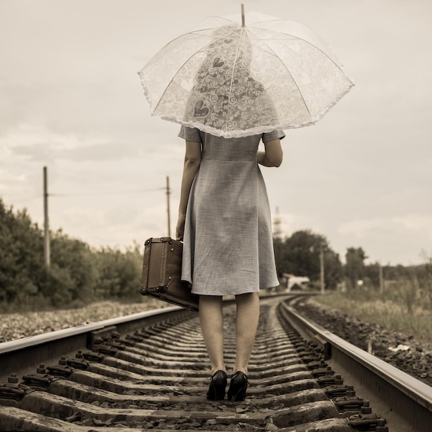 Foto estilo vintage uma garota com um vestido e salto alto com um guarda-chuva e uma mala vai de trem a pé vista traseira sem rosto uma garota retrô caminhando para longe de trem