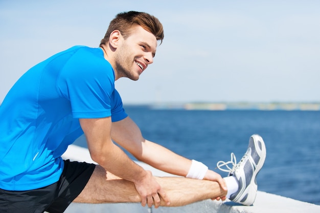 Estirar antes de correr. Apuesto joven haciendo ejercicios de estiramiento y sonriendo mientras está de pie al aire libre