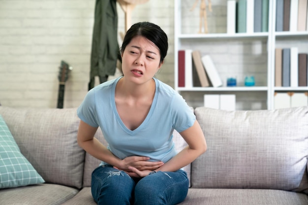 estômago dolorido. mulher chinesa asiática com dor de estômago dolorosa. governanta coreana sofrendo de dor abdominal sentada no sofá cinza conforto na sala de estar em casa com carranca miserável