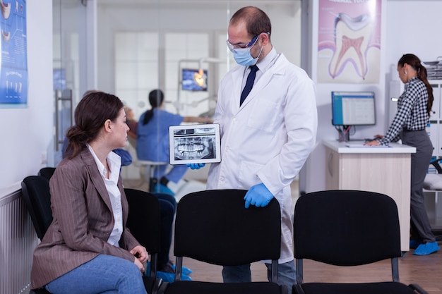 Estomatologista apontando na tela digital explicando a radiografia para uma mulher sentada na cadeira na sala de espera da clínica de estomatologia