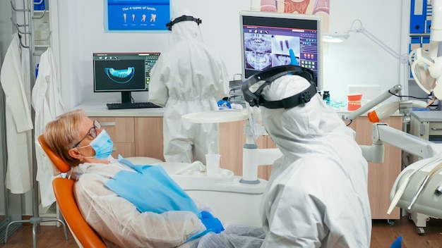 Estomatologista em macacão e paciente olhando para um raio-x dentário digital revisando-o antes da cirurgia durante a pandemia covid-19. Equipe médica usando protetor facial, máscara de proteção e luvas na clínica