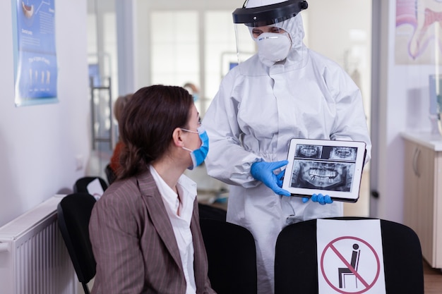 Estomatologista em traje de proteção apontando em um raio-x digital do dente explicando o tratamento do paciente usando comprimido na pandemia covid-19