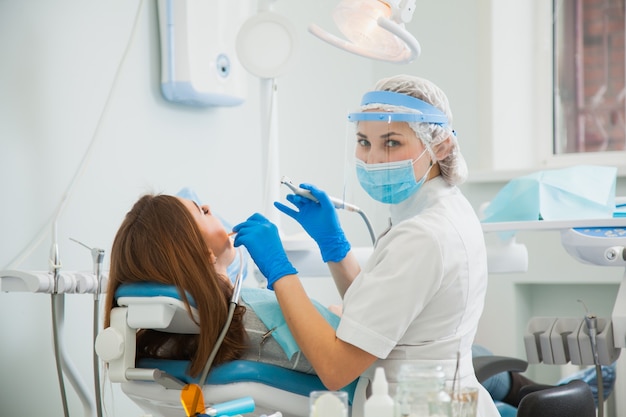 Estomatologista feminina na máscara protetora azul, examinando os dentes do paciente. Tratamento de cárie dentista no consultório da clínica odontológica. Conceito de pessoas, medicina, estomatologia e cuidados de saúde