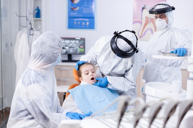 Estomatologista pediátrico fazendo procedimento de higiene oral na menina com babador vestindo terno ppe. Dentista em traje de coronavírus usando espelho curvo durante o exame dos dentes da criança.