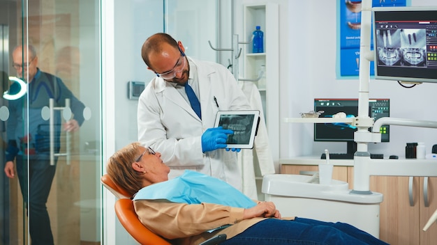 Estomatologista revisando raio x do dente com paciente sênior explicando o tratamento. Dentista mostrando a radiografia dentária de velha usando tablet, médico e enfermeira trabalhando juntos na clínica moderna.