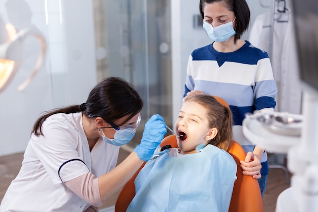 Estomatólogo con espejo en ángulo durante el examen oral de la niña sentada en el sillón dental. Especialista en odontología durante la consulta de cavidades infantiles en el consultorio de estomatología utilizando tecnología moderna.