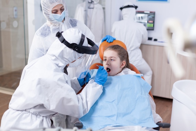 Estomatólogo en traje de ppe durante el examen de los dientes de un niño con babero sentado en el sillón dental. Dentista en traje de coronavirus con espejo curvo durante el examen de los dientes del niño.