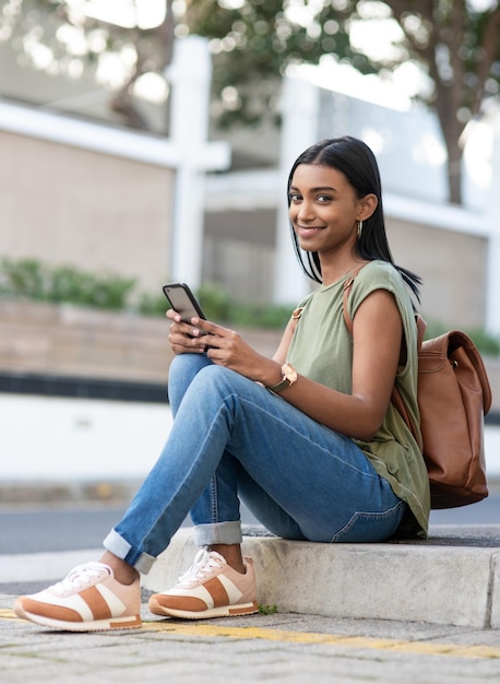 Estou apenas esperando meus amigos se juntarem a mim retrato de corpo inteiro de uma jovem atraente usando um smartphone enquanto está sentado na cidade