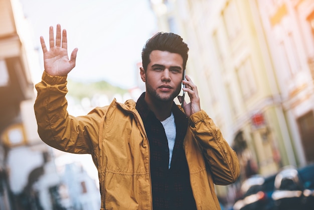 Estou bem aqui! Jovem bonito falando no telefone inteligente e acenando para alguém em pé na rua da cidade