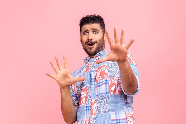 Estou com medo. Retrato de homem barbudo assustado chocado na camisa azul estilo casual gritando de horror e medo, mantendo as mãos levantadas para se defender. Tiro de estúdio interior isolado no fundo rosa.