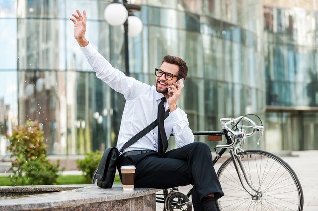 ¡Estoy aquí! Alegre joven empresario hablando por el teléfono móvil y saludando a alguien mientras está sentado cerca de su bicicleta con edificio de oficinas en el fondo