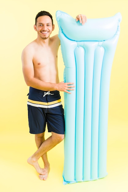 Estoy listo para relajarme en la piscina. Joven hispano en traje de baño que lleva un colchón inflable para descansar en la playa