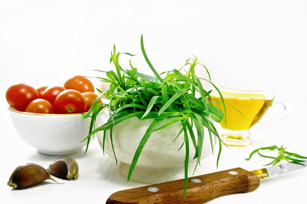 Estragón fresco en un mortero tomates y champiñones en tazones aceite vegetal en salsera dientes de ajo y un cuchillo sobre fondo de tablero de madera clara