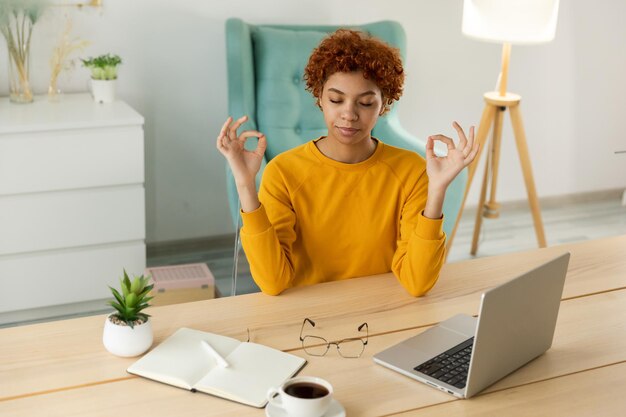 Sin estrés, mantén la calma Empresaria africana consciente practica ejercicios de respiración en la oficina en casa Mujer joven pacífica en el lugar de trabajo disfruta del yoga ojos cerrados manos en el gesto de mudra de la barbilla Meditación en la oficina