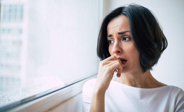 Estrés profundo. Foto de primer plano de una mujer triste que mira por la ventana y se muerde las uñas con angustia.