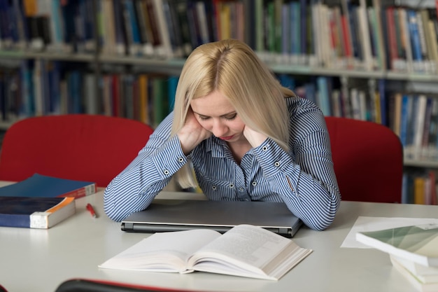 Estressada jovem estudante lendo livro enquanto está sentado na biblioteca