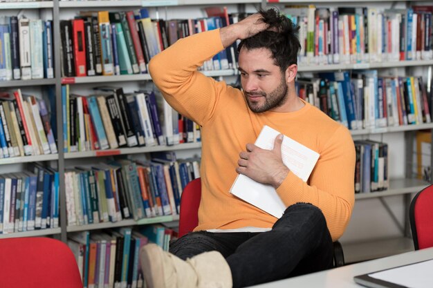 Estressado jovem estudante lendo livro enquanto sentado na biblioteca