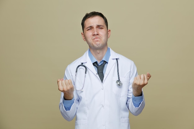 Estressado jovem médico masculino vestindo roupão médico e estetoscópio no pescoço, olhando para a câmera, mantendo os punhos no ar isolados no fundo verde-oliva