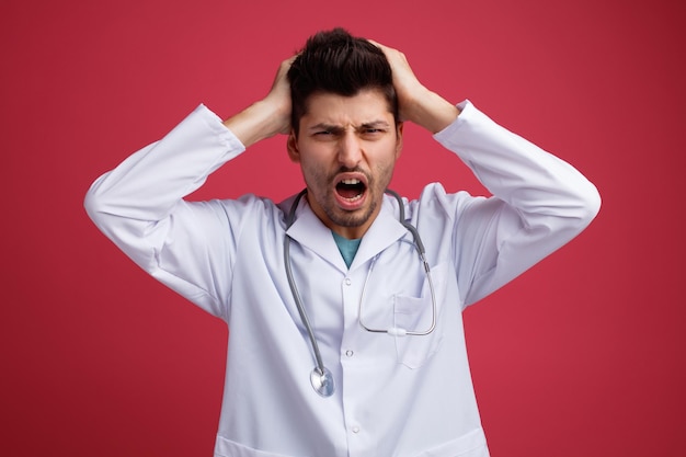 Estressado jovem médico masculino vestindo uniforme médico e estetoscópio em volta do pescoço, mantendo as mãos na cabeça isolada em fundo vermelho