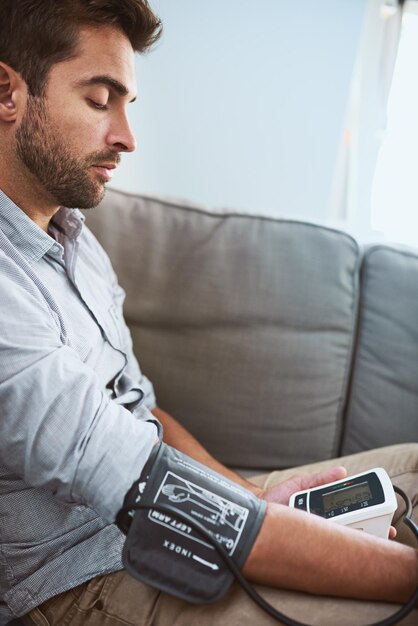 Foto estresse hipertensivo e homem testam pressão arterial em um sofá doméstico ou sala de estar para exame médico ou de saúde monitoram leitura e homem com equipamento ou máquina para doenças crônicas em uma casa