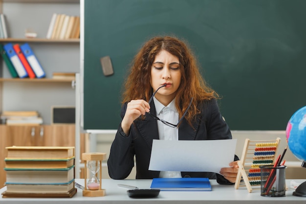 estricta joven maestra con gafas sosteniendo y leyendo papel sentada en el escritorio con herramientas escolares en el aula