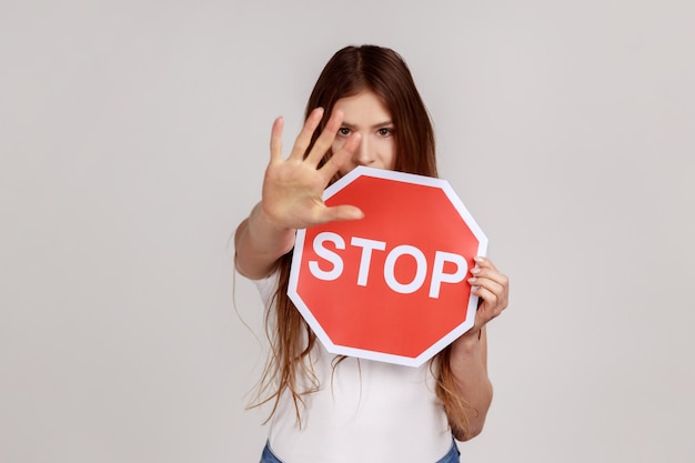 Estricta mujer mandona que mira enojada y muestra un gesto de alto, sosteniendo una señal de tráfico como advertencia de prohibición, acceso prohibido, usando una camiseta blanca. Disparo de estudio interior aislado sobre fondo gris.