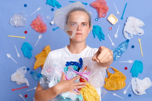 Estricta mujer seria enojada con camisa blanca aislada en la pared azul con basura alrededor limpiando basura apuntando a la cámara pidiéndole que no tire basura