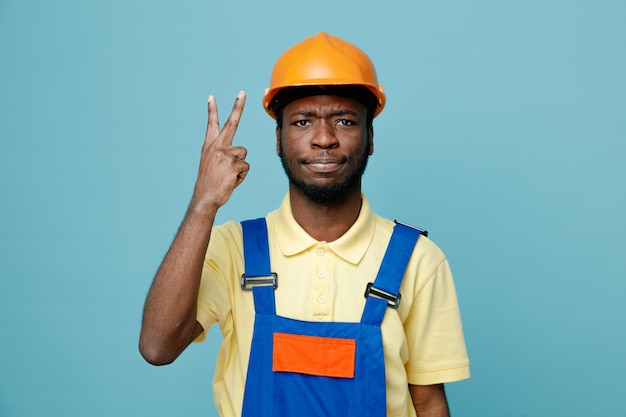 Estricto gesto de paz mostrando joven constructor afroamericano en uniforme aislado sobre fondo azul.