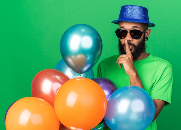 Estricto joven afroamericano con sombrero de fiesta y gafas sosteniendo globos mostrando gesto de sí aislado en la pared verde