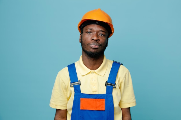 Estricto joven constructor afroamericano en uniforme aislado sobre fondo azul.