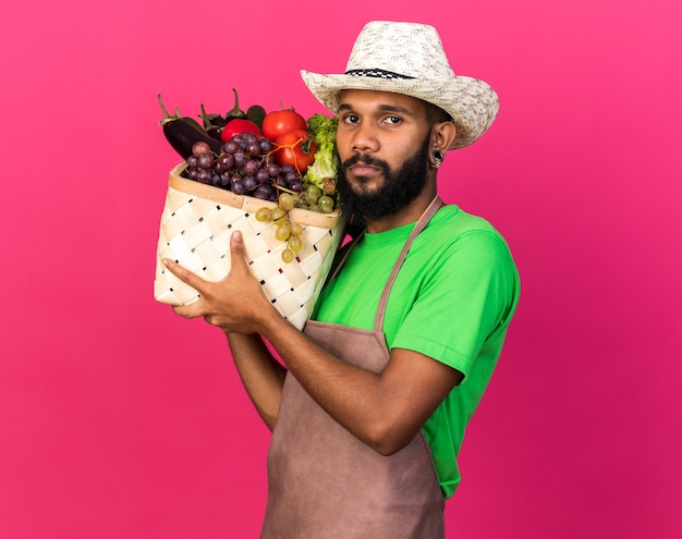 Estricto joven jardinero afroamericano vistiendo sombrero de jardinería sosteniendo canasta de verduras