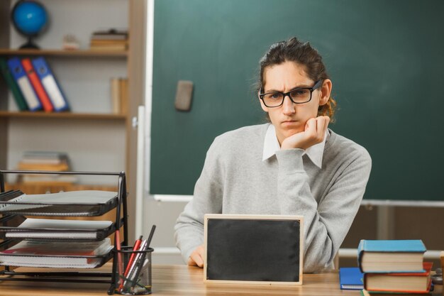 estricto poner la mano en la barbilla joven maestro con gafas sentado en el escritorio sosteniendo una mini pizarra con herramientas escolares en el aula