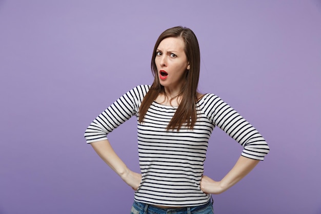 Estrita irritada morena jovem em roupas listradas casuais posando isolado no fundo roxo violeta. Conceito de estilo de vida de pessoas. Mock up espaço de cópia. De pé com os braços cruzados na cintura, praguejando.