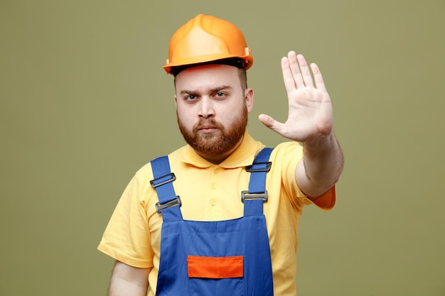 Estrito mostrando gesto de parada jovem construtor de uniforme isolado em fundo verde