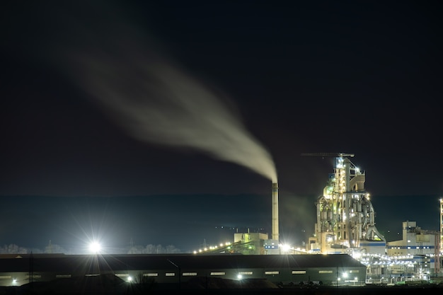 Estrutura de concreto alta da fábrica de cimento com guindaste de torre e chaminé de fumo à noite.