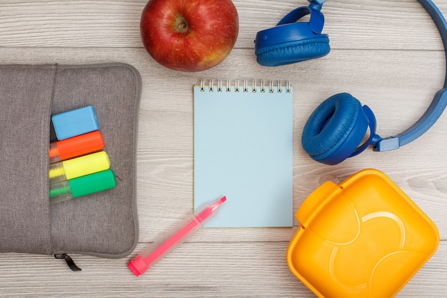 Estuche bolso-lápiz con rotuladores de colores y rotulador, manzana, cuaderno azul, auriculares y fiambrera sobre fondo de madera gris. Vista superior con espacio de copia. Concepto de regreso a la escuela. Suministros escolares.