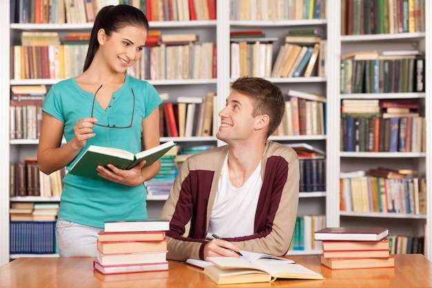 Estudando juntos. Jovem alegre sentado à mesa enquanto uma bela mulher em pé perto dele segurando um livro