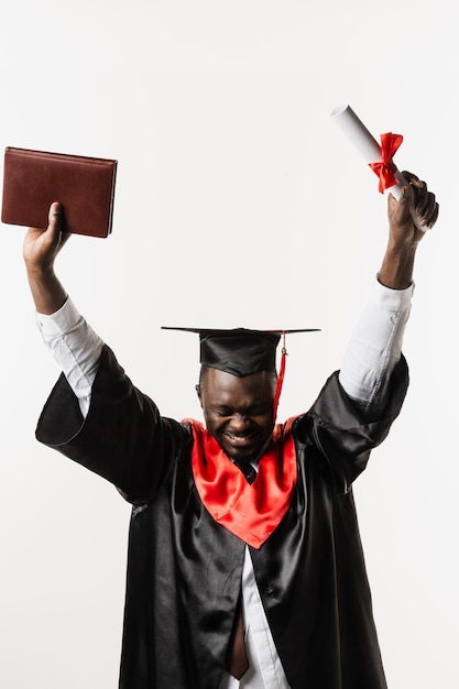 Estudante africano feliz em vestido de formatura preto e boné levanta diploma de mestrado acima da cabeça em fundo branco Homem africano graduado está se formando na faculdade e comemorando conquistas acadêmicas