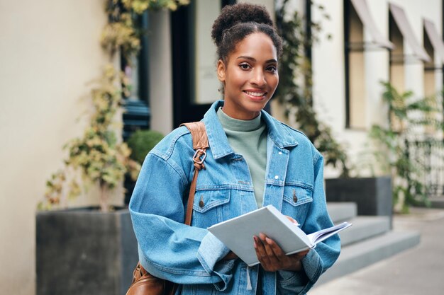 Foto estudante afro-americana bastante casual em jaqueta jeans com mochila e livro olhando alegremente para a câmera na rua da cidade