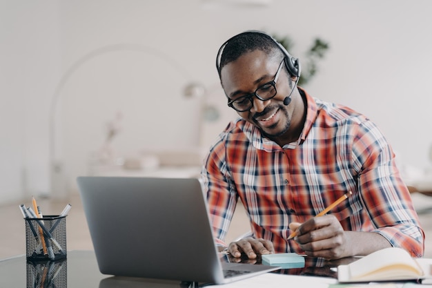 Estudante afro-americano em aprendizado de fone de ouvido no laptop on-line faz anotações Educação à distância