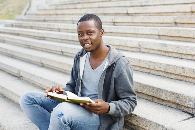 Estudante afro-americano lendo livro nas escadas da universidade, preparando-se para os exames na faculdade ou universidade a sorrir. conceito de educação, espaço de cópia