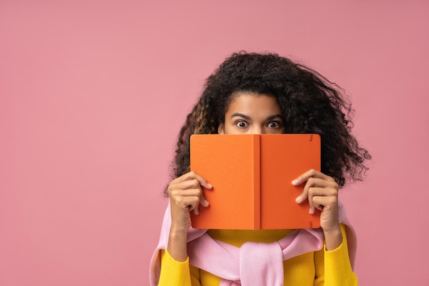 Estudante afro-americano segurando livro perto do rosto isolado no espaço de cópia de fundo rosa Educação