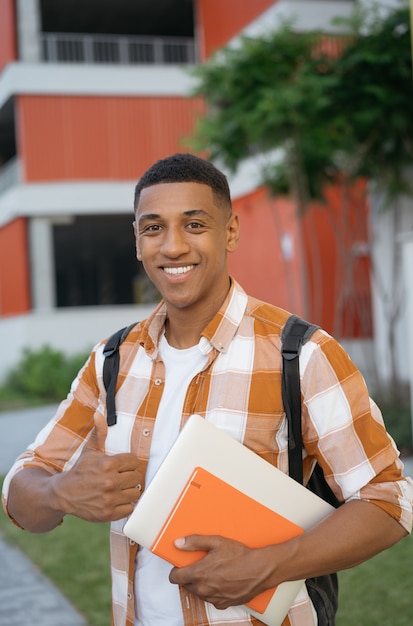 Foto estudante afro-americano sorridente, segurando o polegar, olhando para a câmera em pé no campus da universidade