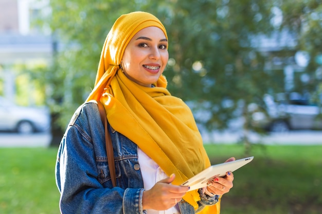 Estudante árabe. Bela aluna muçulmana usando o tablet de exploração do hijab amarelo brilhante.